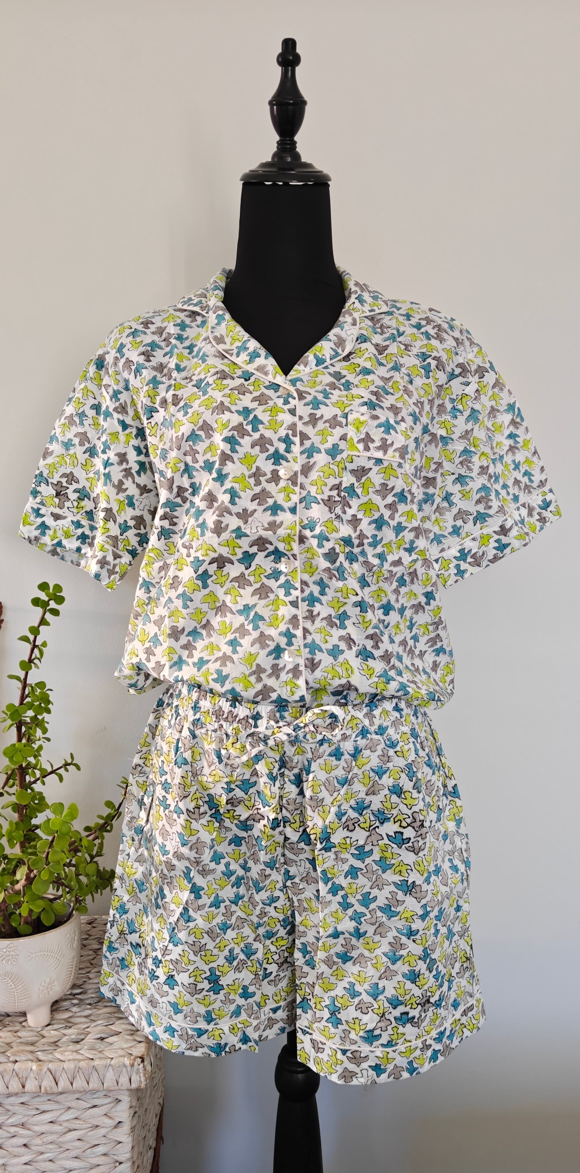 A two-piece matching set featuring a short-sleeve V-neck top and coordinating shorts, both decorated with a colorful floral print in blue, green, yellow, and purple on a white birds background The set is displayed on a black mannequin torso next to a wicker basket with potted plants.