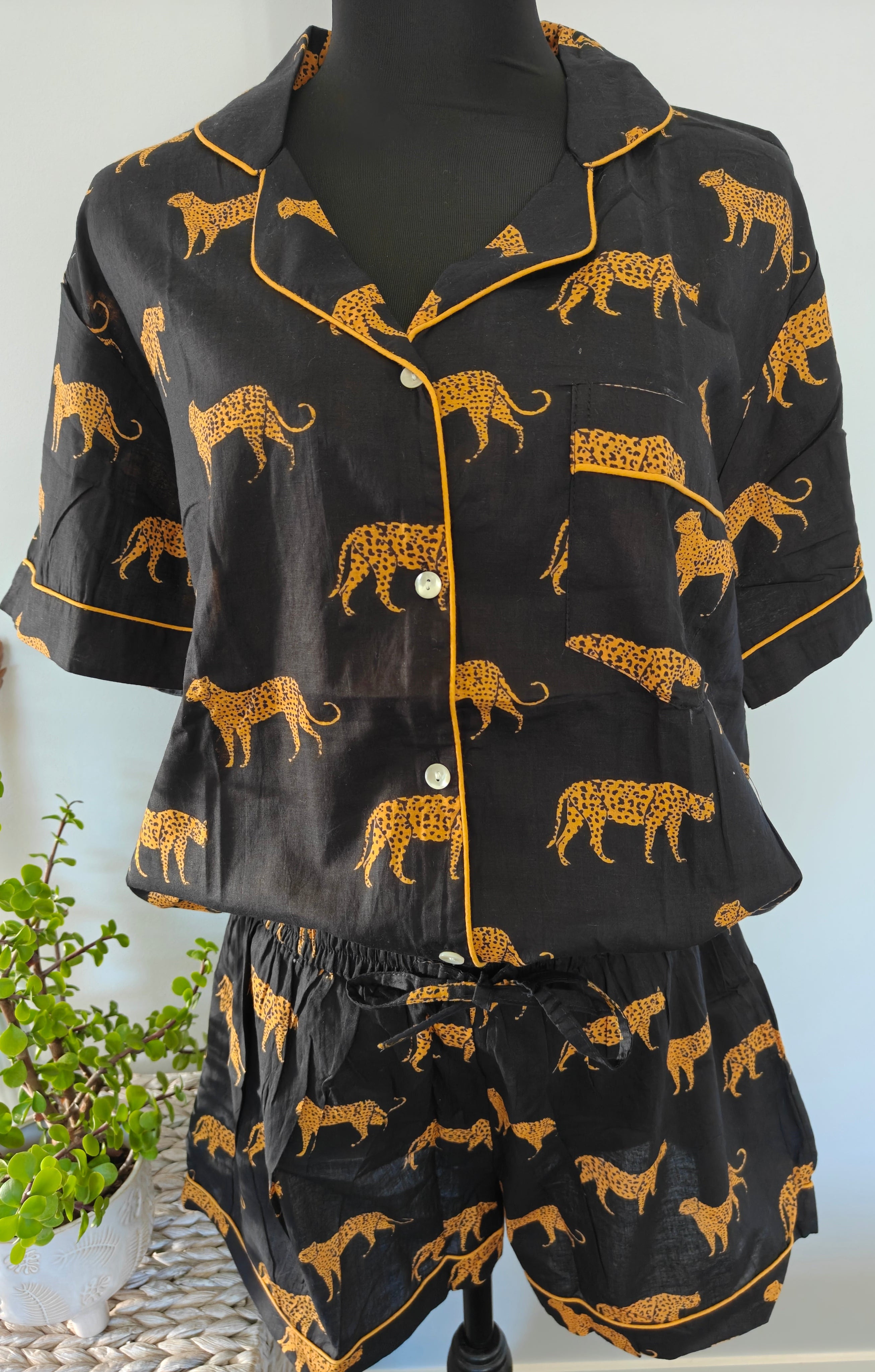 A two-piece pyjama set displayed on a black mannequin torso, featuring a short-sleeve button-front top and matching shorts in black with a mustard yellow leopard print and piping.