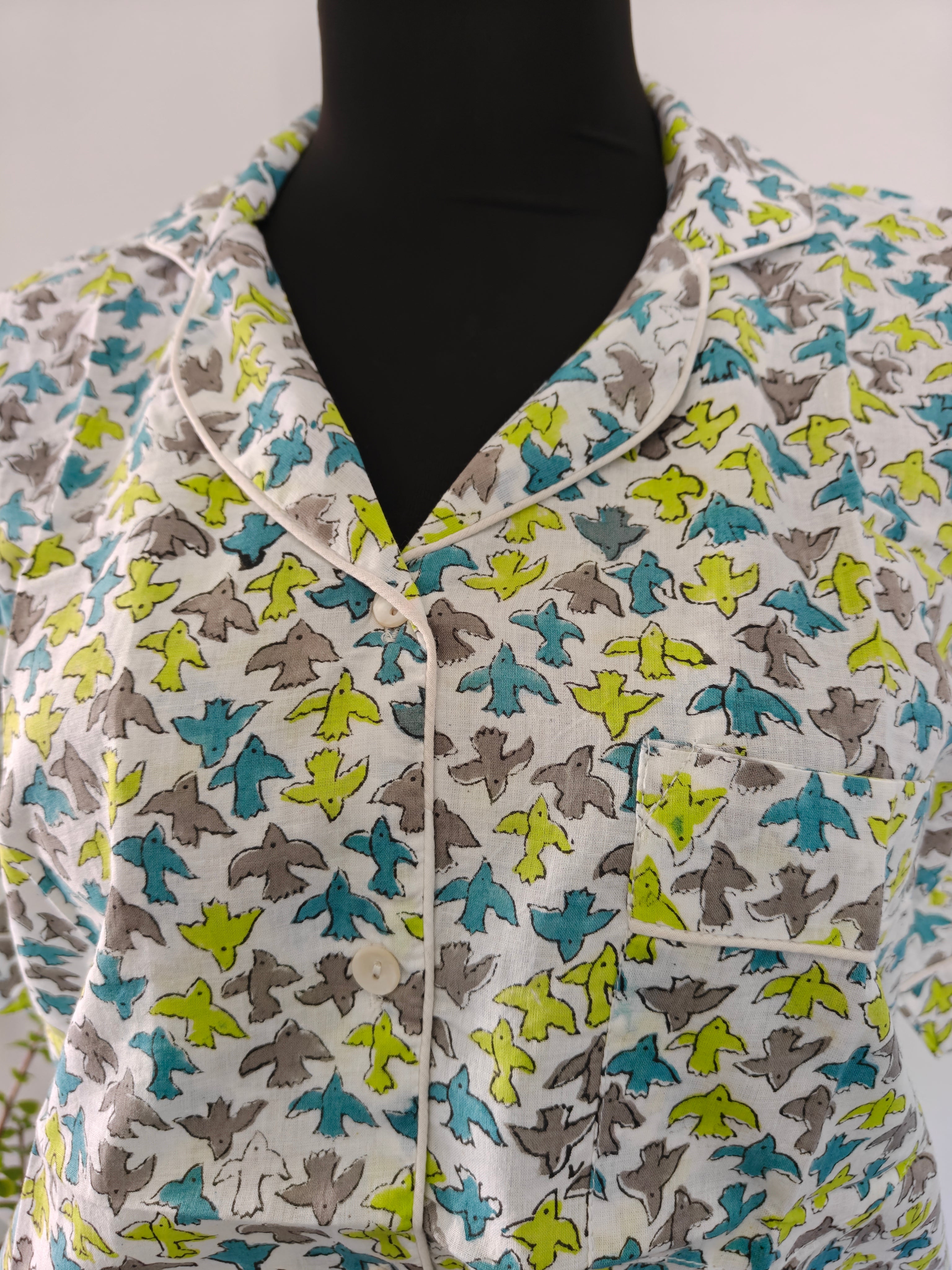 A close-up of a vintage-style button-up pj set with a white base and an all-over pattern of small birds in teal, lime green, and gray. 