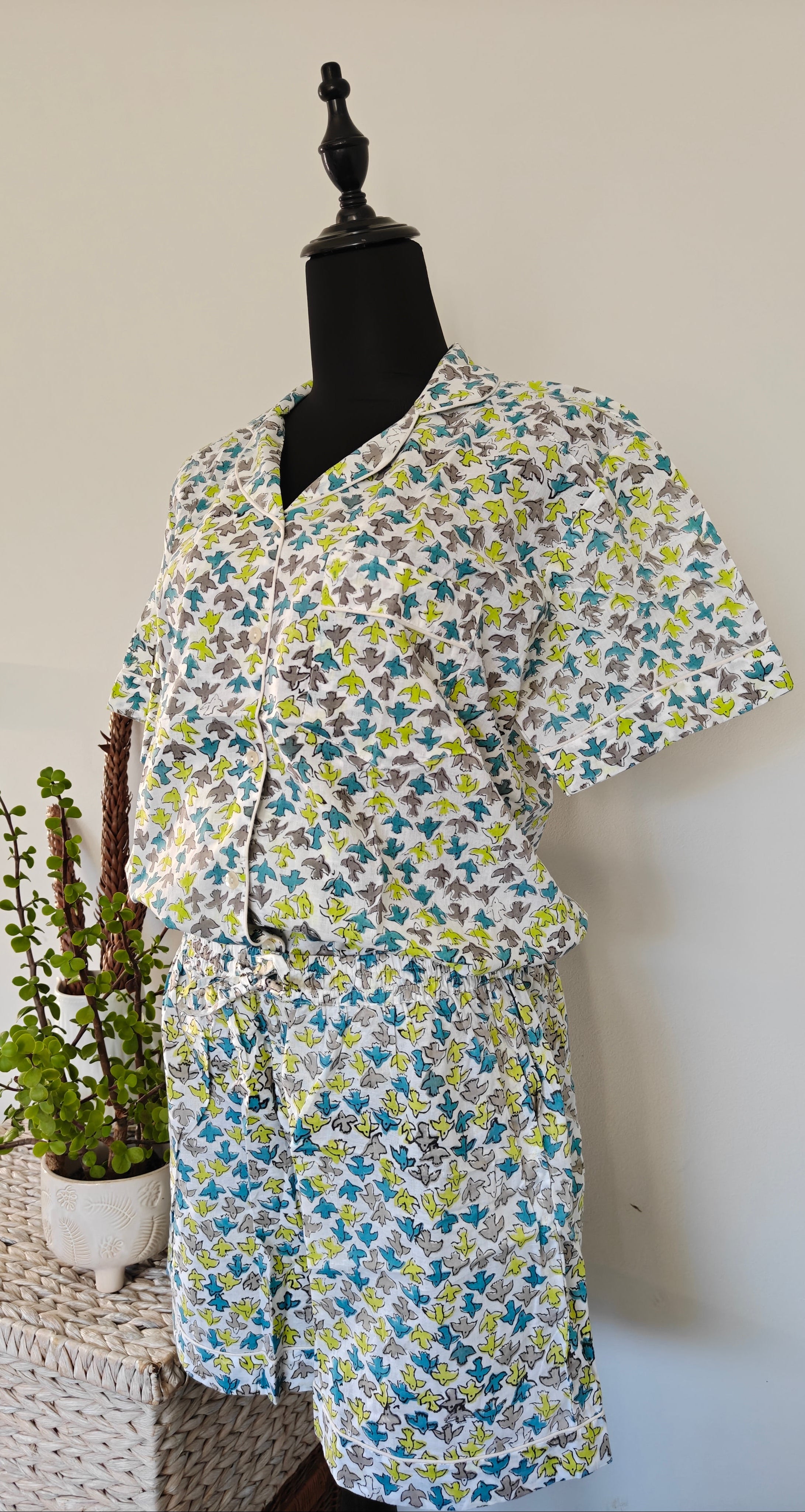 A short-sleeve pyjama top and matching shorts set displayed. The set features a busy, dense floral pattern in shades of blue, green, yellow, and gray on a white background. Birds print