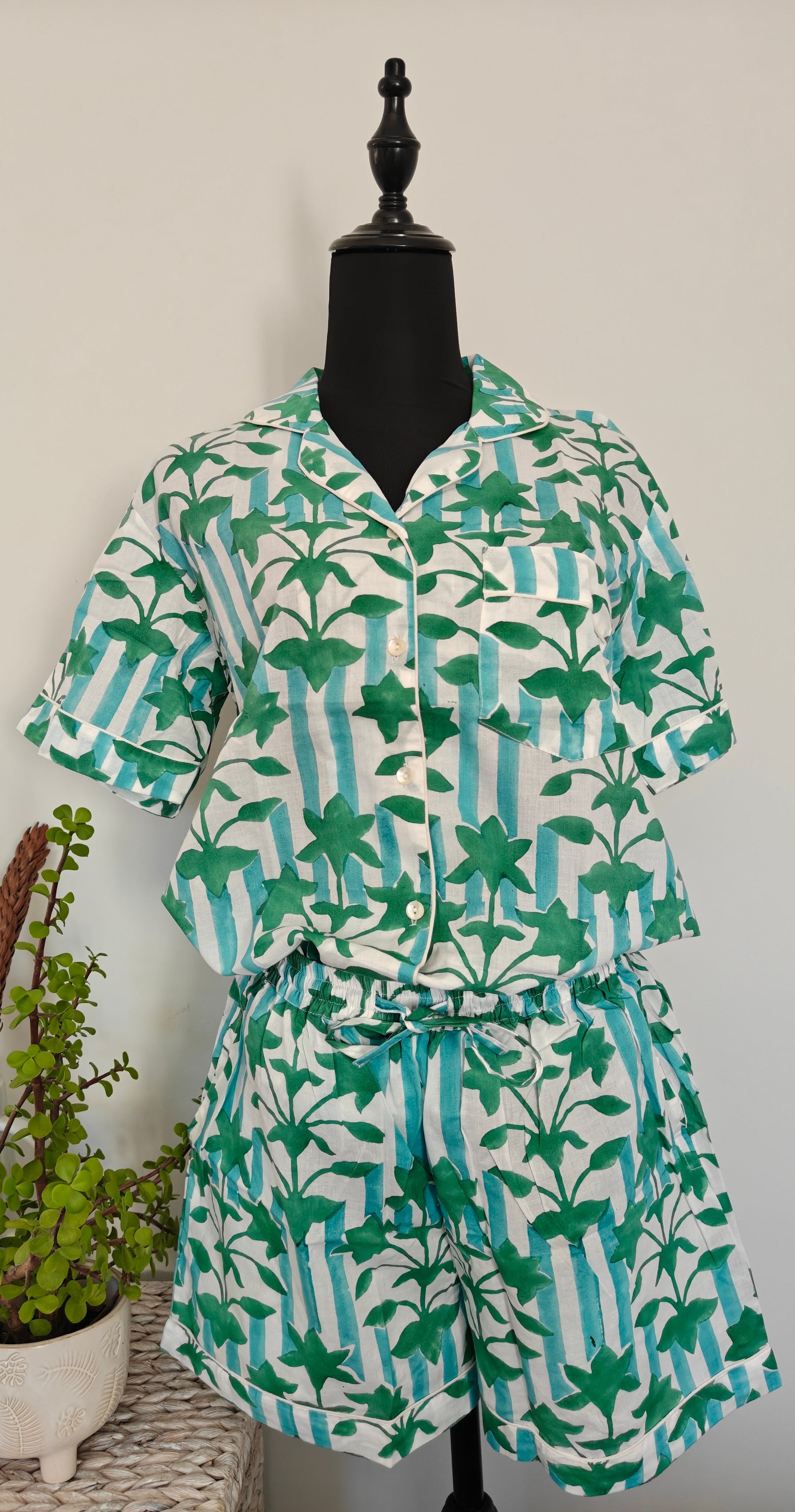 A two-piece matching set consisting of a short-sleeve button-up shirt and shorts, displayed on a black mannequin. The set features a white base with vertical light blue stripes and green tropical palm leaf prints.