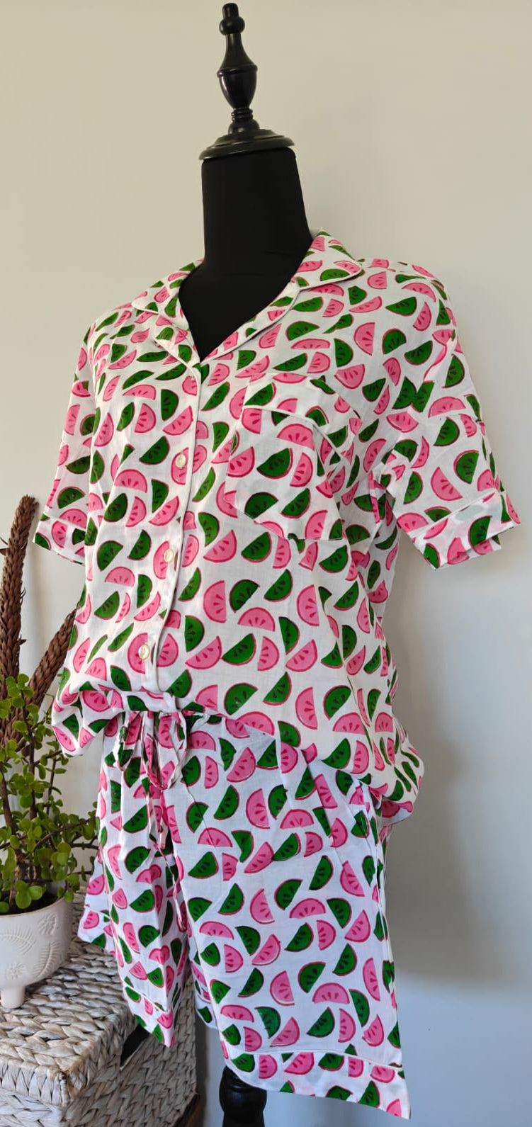 A short-sleeve white shirt dress with a pink watermelon and green seed print, displayed on a black mannequin torso next to a potted plant on a wicker table.