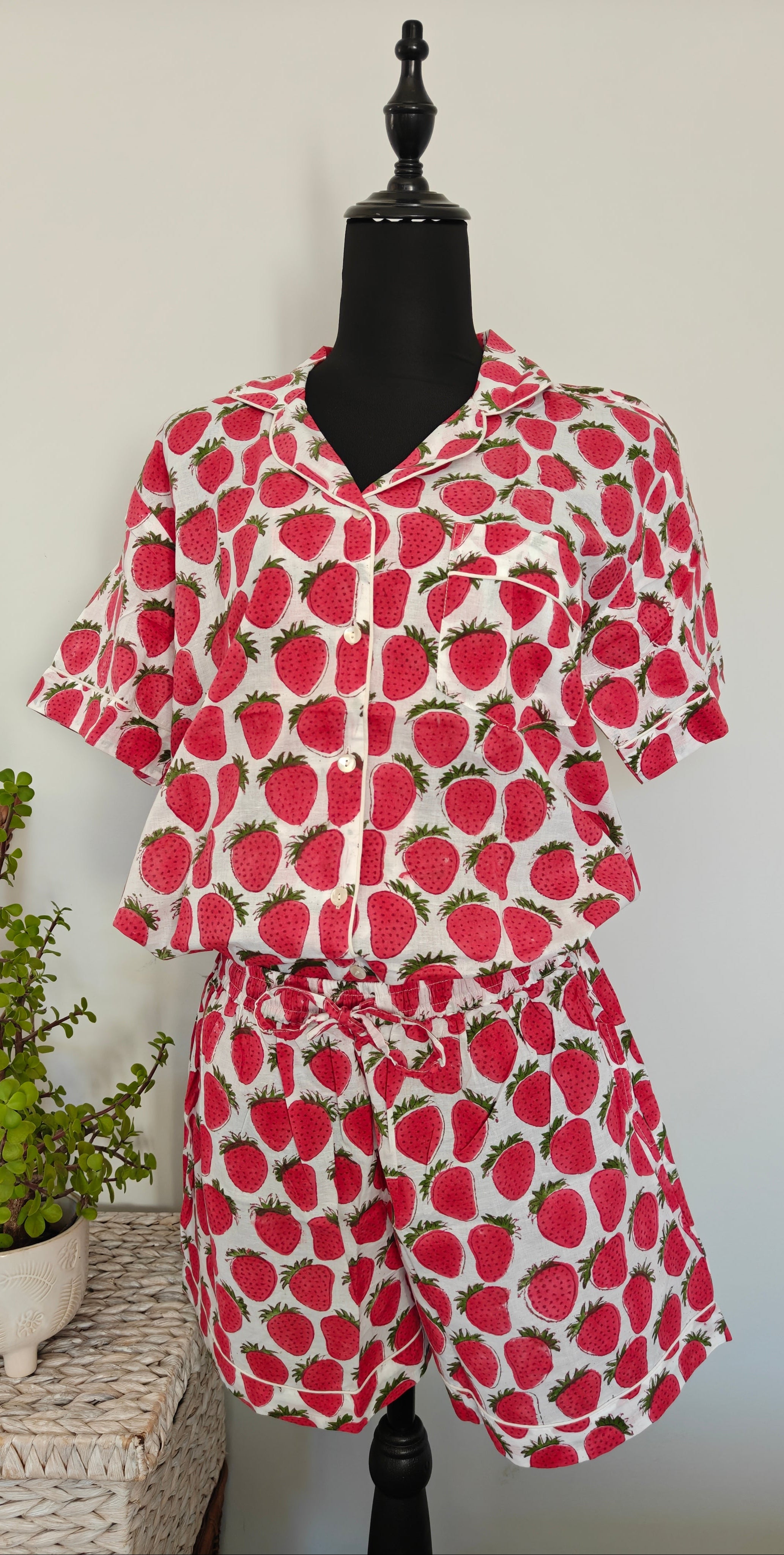 A two-piece matching set consisting of a short-sleeve button-up shirt and shorts, featuring a red strawberry print on a white background, displayed on a black mannequin torso with a small potted plant on a woven side table.