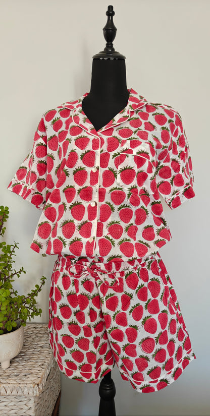 A two-piece matching set consisting of a short-sleeve button-up shirt and shorts, featuring a red strawberry print on a white background, displayed on a black mannequin torso with a small potted plant on a woven side table.