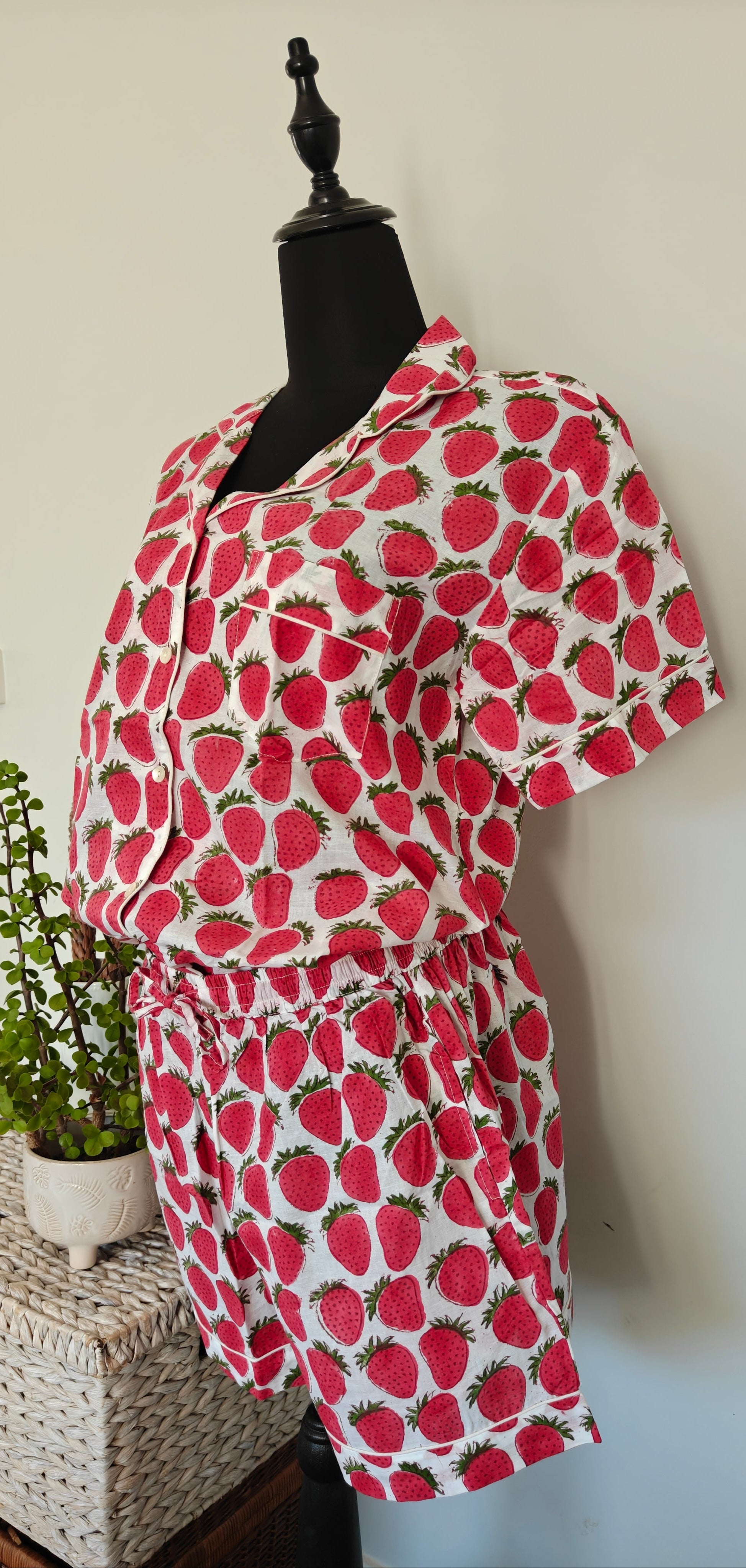 A strawberry-print short-sleeve romper displayed on a black dress form mannequin indoors, next to a small potted green plant on a woven storage box against a plain light wall.